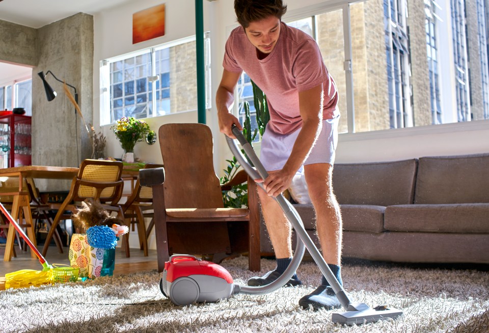 A young man vacuuming the carpet in a sunlit living room.