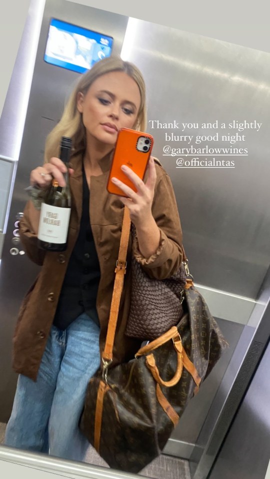 Emily Atack holding a bottle of wine and taking a selfie in an elevator.