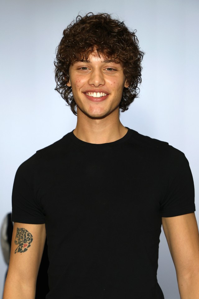 Bobby Brazier smiles, wearing a black t-shirt with a leopard tattoo on his right bicep at London Fashion Week.