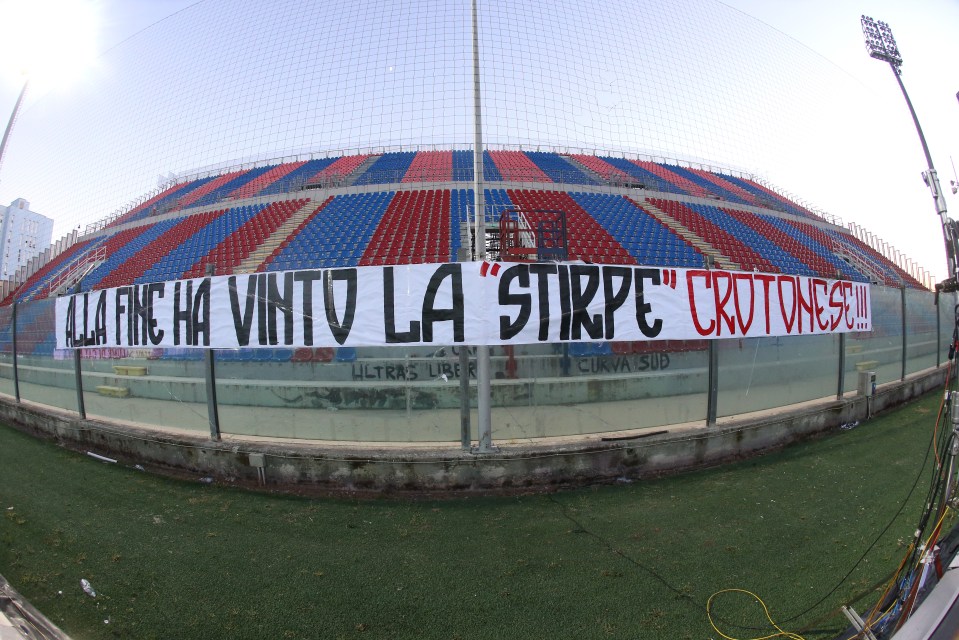 An empty soccer stadium with red and blue seats and a banner that reads "ALLA FINE HA VINTO LA “STIRPE” CROTONESE!!!"