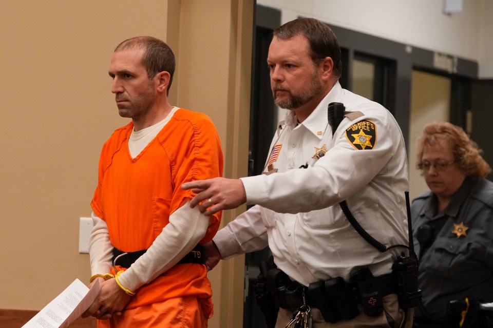 A man in an orange jumpsuit is escorted by a sheriff's deputy in a courtroom.