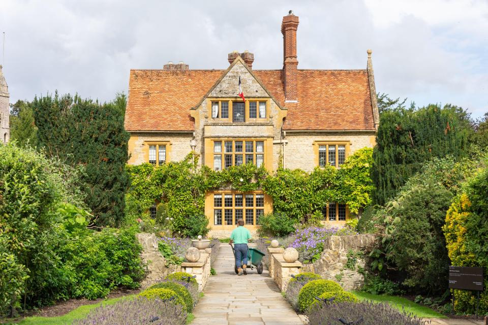 Gardener wheeling a wheelbarrow down a stone path towards a 15th-century manor house.