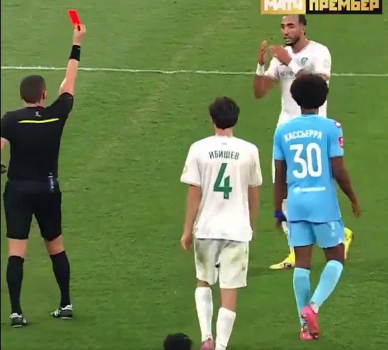 A soccer referee holds up a red card, indicating an ejection, while standing on a field with players.