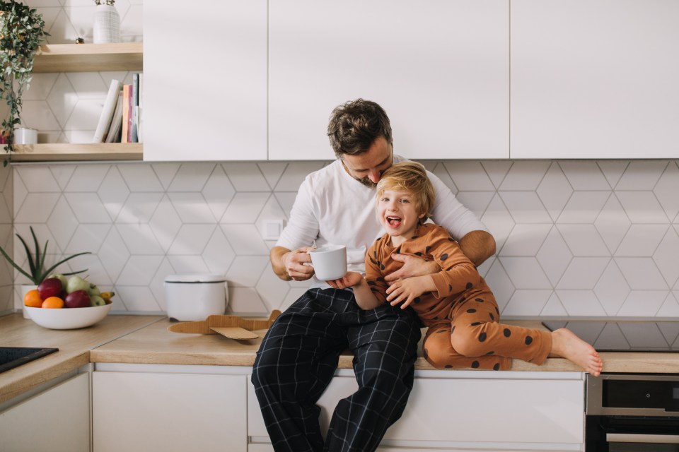 Father embracing his son in pajamas, who is sitting on a kitchen counter and smiling at the camera.