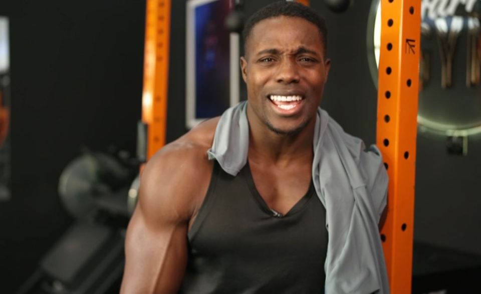 A muscular man in a black tank top with a gray towel over his shoulder, smiling with his teeth showing.