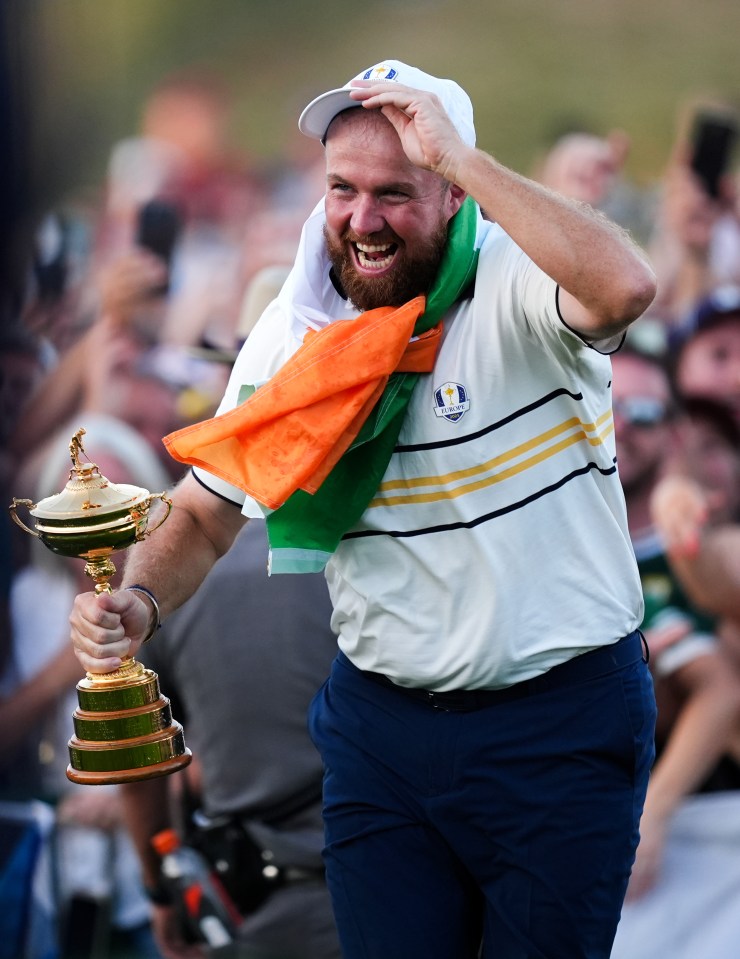 Shane Lowry celebrating with the Ryder Cup Trophy, with an Irish flag draped over his shoulder.