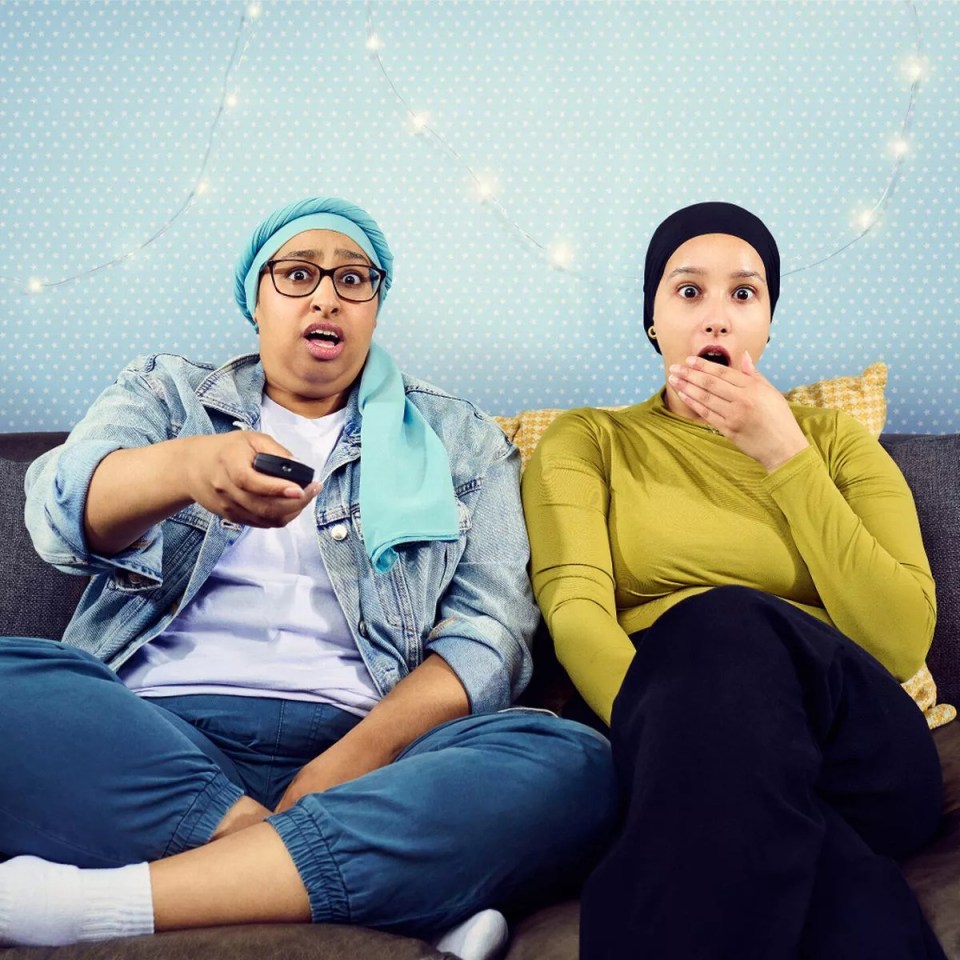 Two young women with head coverings look shocked while watching TV.