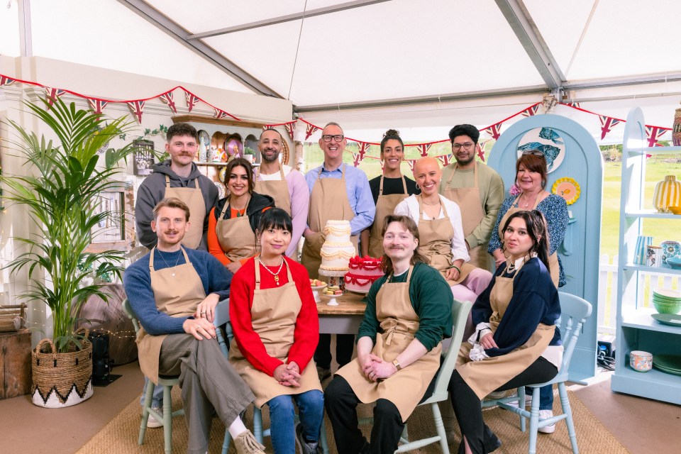 Contestants of The Great British Bake Off Series 16, Toby, Nadia, Aaron, Leighton, Jessika, Jasmine, Hassan, Lesley, Tom, Pui Man, Iain, and Nataliia, posing together in the Bake Off tent.