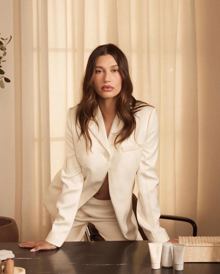 Hailey Bieber leaning on a table with Rhode skincare products.
