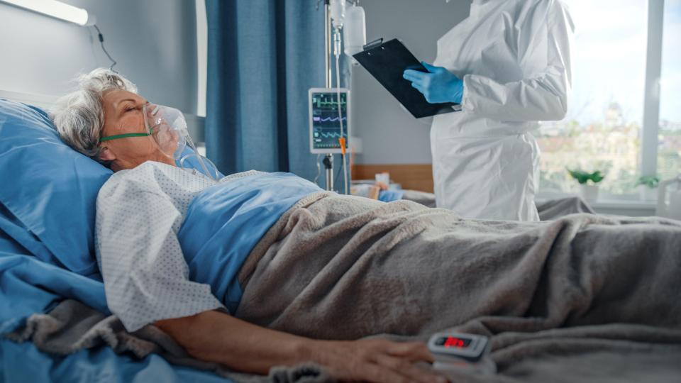 Doctor wearing a coverall writes down vitals of a senior female patient with an oxygen mask resting in bed.