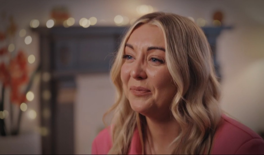 A woman with blonde hair, in a pink top, looking emotional.