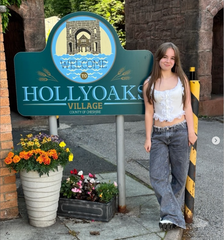 A girl smiling in front of a “Welcome to Hollyoaks Village” sign.
