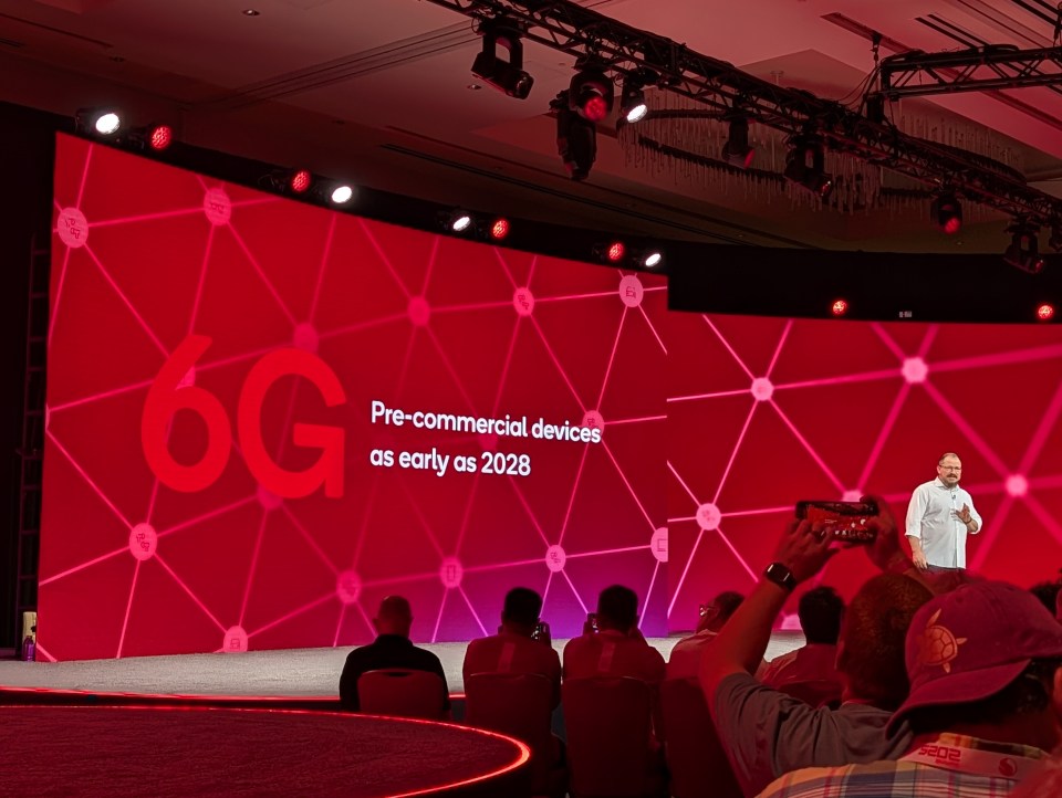A speaker on stage with a red screen behind him displaying "6G Pre-commercial devices as early as 2028".