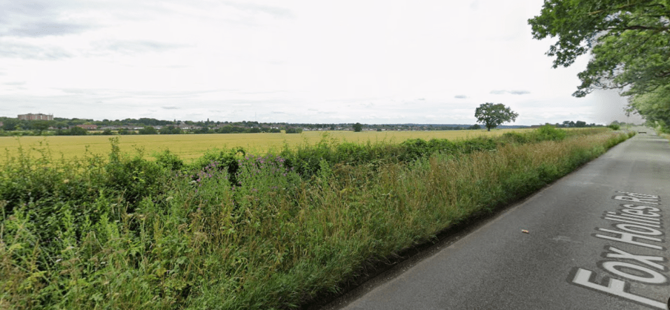 A paved road with "Fox Hollies Rd" painted on it, running alongside a field with a town in the distance.