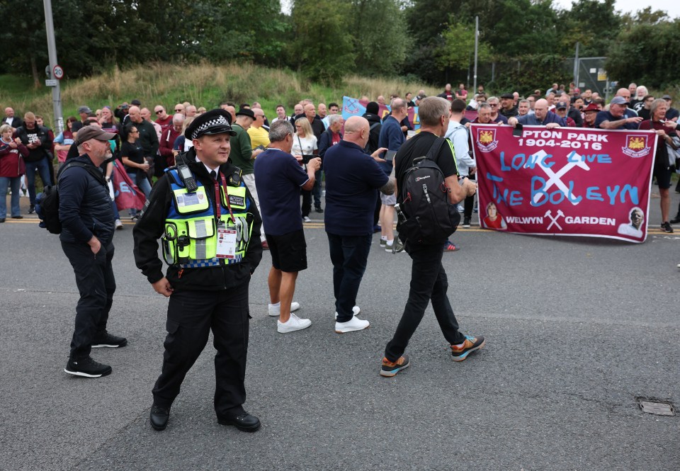 A police officer in uniform stands on a road with West Ham United fans protesting in the background, holding a banner that says "Upton Park 1904-2016 Long Live The Boleyn".