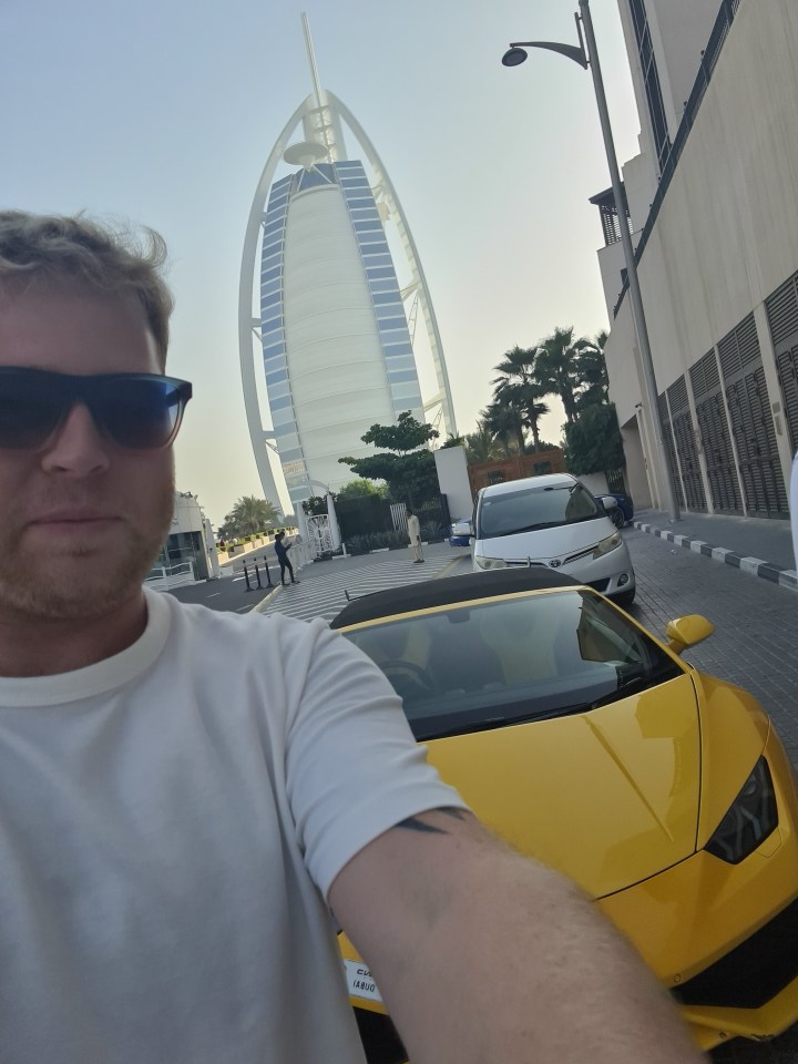 Man taking a selfie in front of a yellow Lamborghini with the Burj Al Arab in the background.