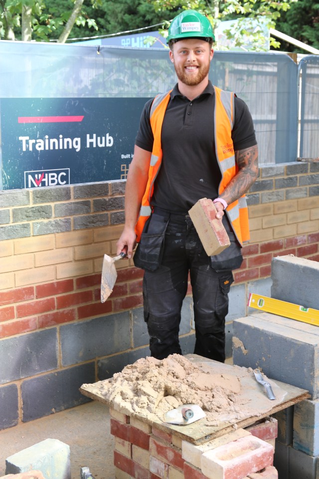 Toby Egan, a male wearing a construction helmet, polo shirt, and safety vest, holding a brick and a trowel, standing in front of a "Training Hub" sign.