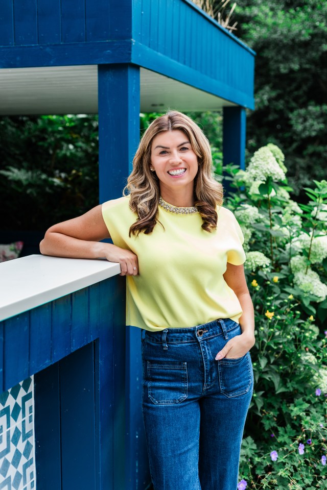 A smiling woman with light brown hair in a yellow top and jeans leaning on a blue railing.
