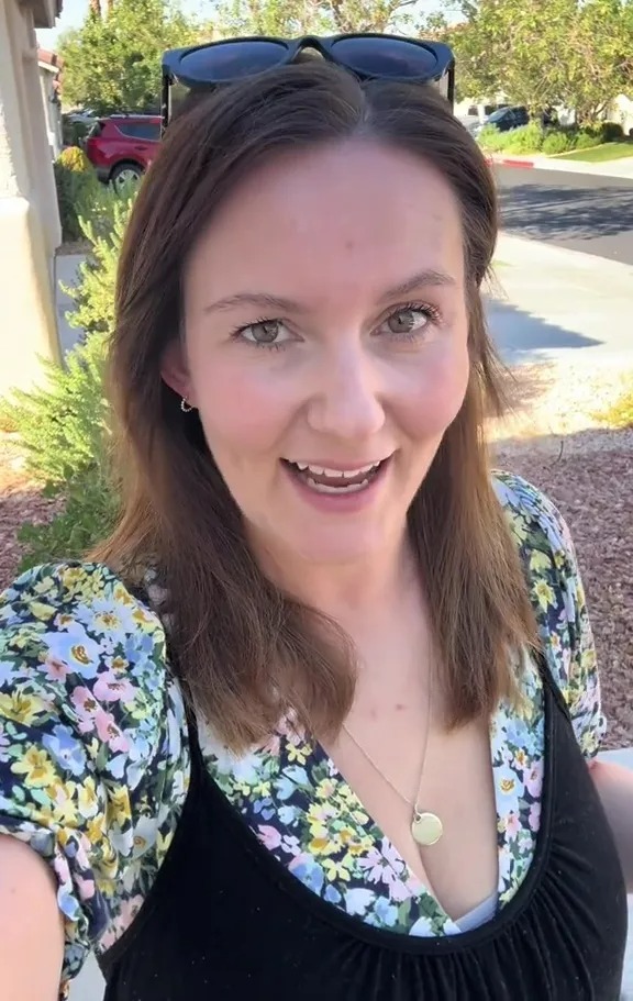 A smiling woman in a floral top and black tank top standing outdoors on a sunny day.