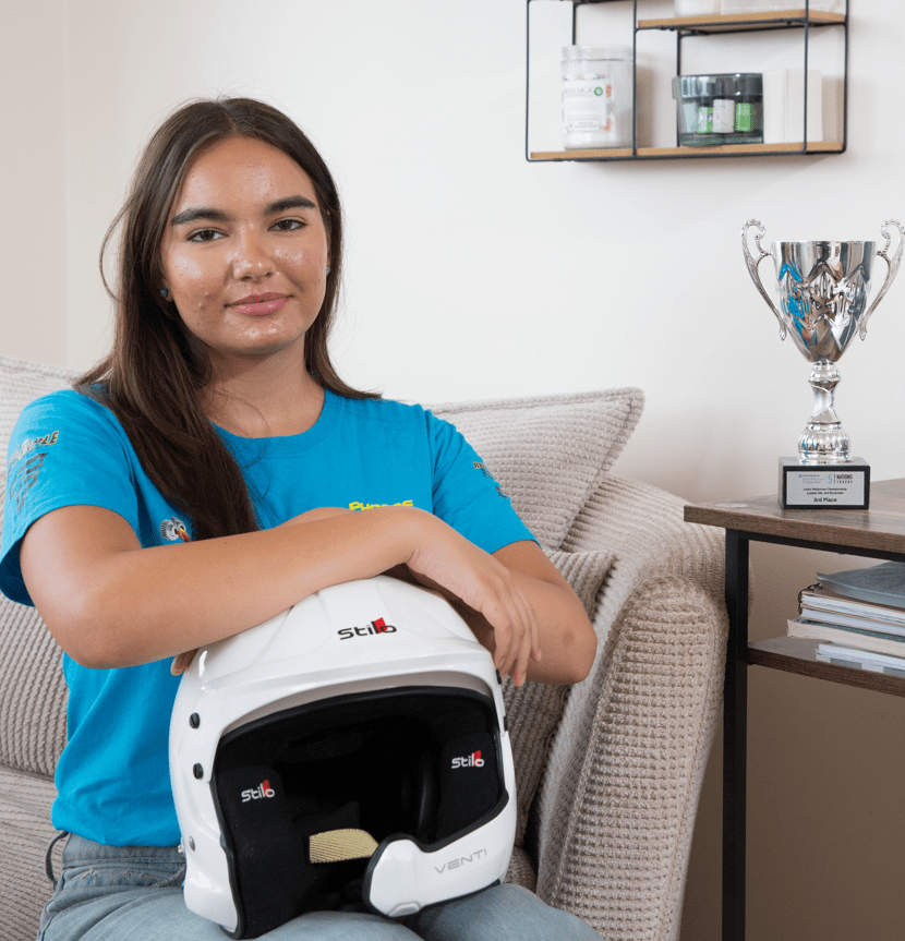 Teenage girl holding racing helmet, sitting on couch near trophy.