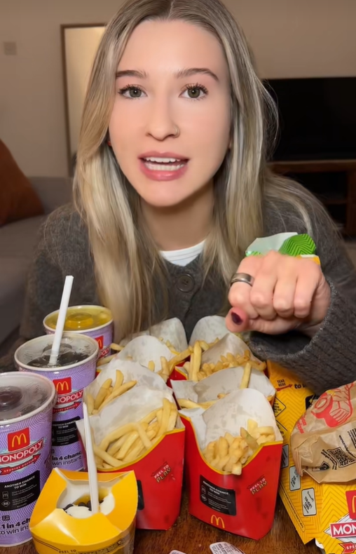 A young woman sits behind several containers of McDonald's food, holding up a McDonald's Monopoly peel-off game piece.