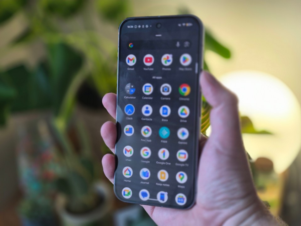 A hand holding a smartphone displaying the app drawer with various Google apps.
