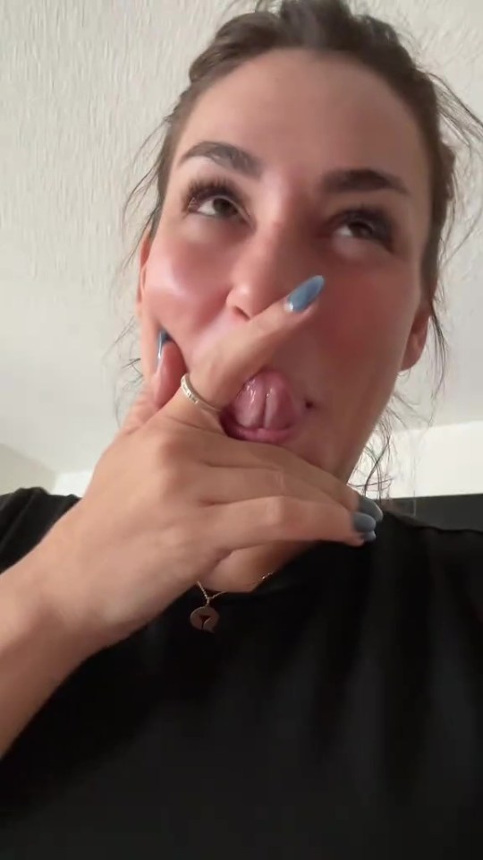 A woman with blue nail polish and a ring on her finger holds her mouth open with her fingers, showing her tongue.