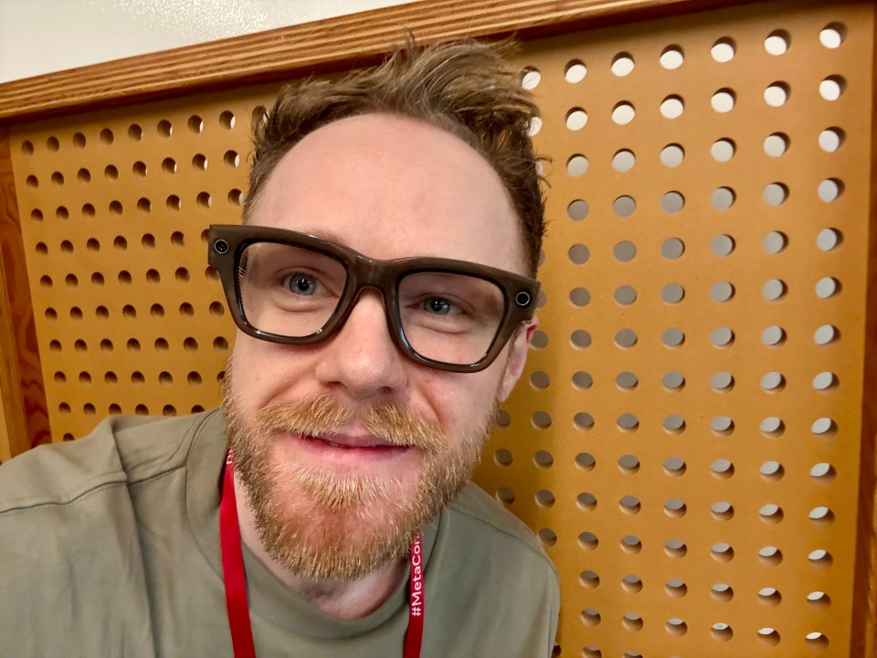 A man wearing thick-framed smart glasses, an olive green shirt, and a red #MetaCon lanyard, smiles.