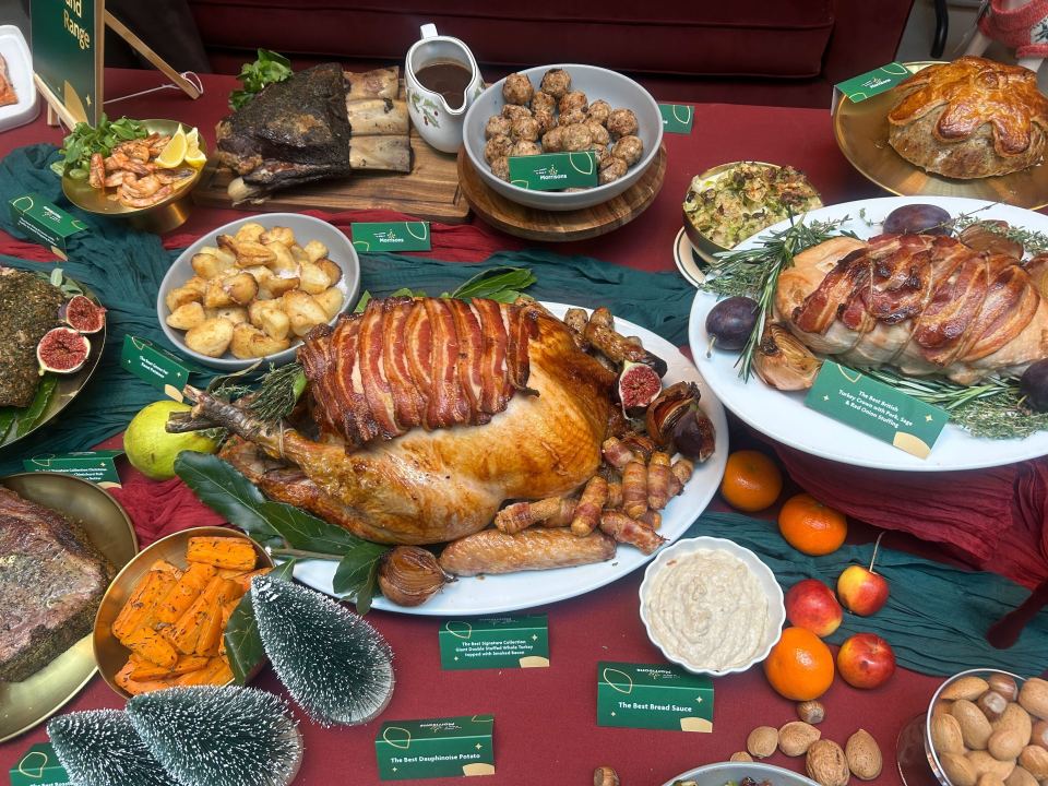 A festive spread of cooked meats including two turkeys, roasted potatoes, vegetables, a gravy boat, and nuts on red and green cloths.