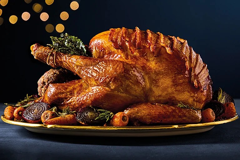 A golden roasted whole turkey on a platter with roasted vegetables and festive lights in the background.