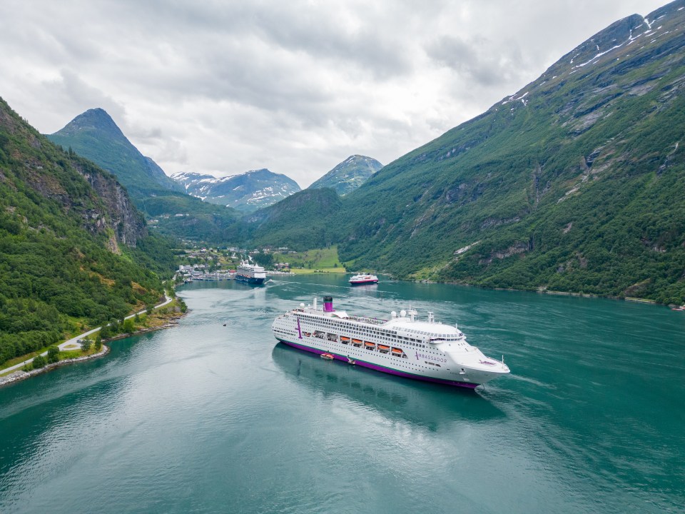 A cruise ship, the Ambience, sailing through a fjord with mountains on either side.