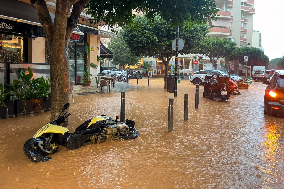 Flooded street in Ibiza, Spain with an overturned yellow scooter and vehicles partially submerged in brown floodwaters.