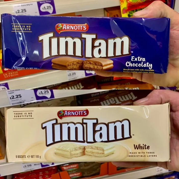 Hands holding a dark blue Arnott's Tim Tam "Extra Chocolaty" package and a white Arnott's Tim Tam "White" package.