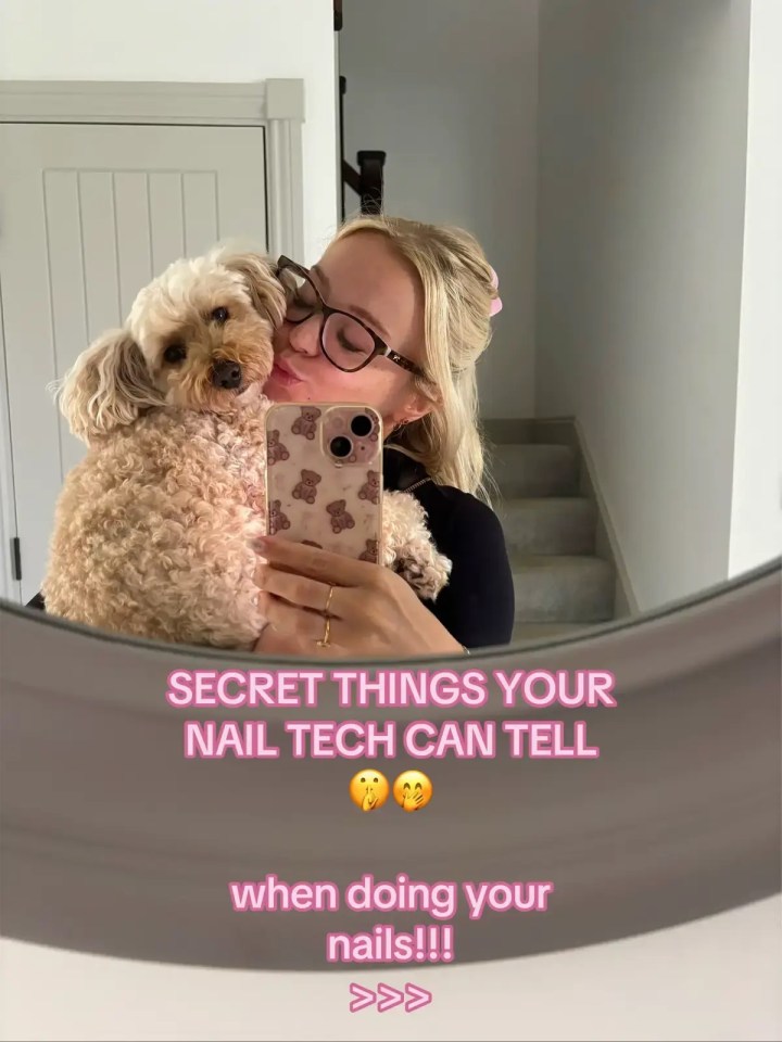 A blonde woman in glasses kisses a small curly-haired dog while holding up a phone with a teddy bear case, reflected in a mirror with pink text overlay.