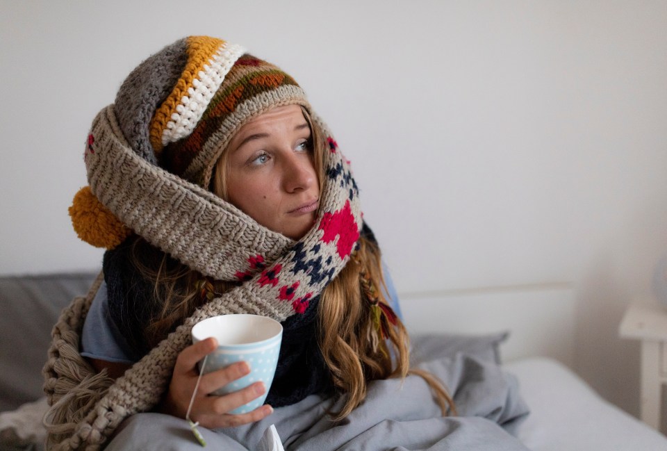 Young woman in knit hat and scarf holding a cup of tea, looking displeased.