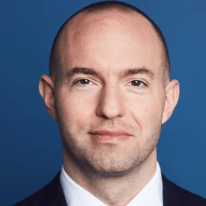 Headshot of a man with a shaved head and a light beard, wearing a suit and smiling.