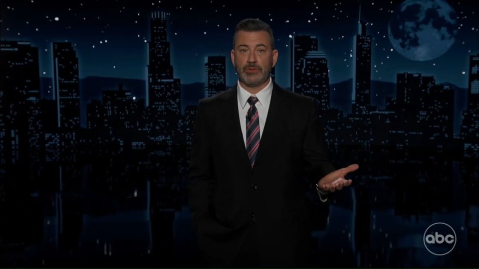 Jimmy Kimmel on stage in a suit with a city skyline and moon backdrop.