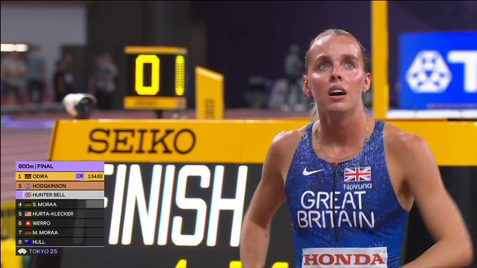 A British female athlete in a blue tank top looking up, with a scoreboard displaying race results behind her.
