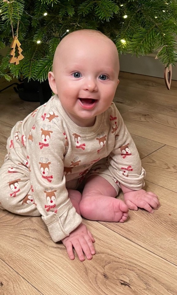 A baby boy in reindeer pajamas sitting in front of a Christmas tree.