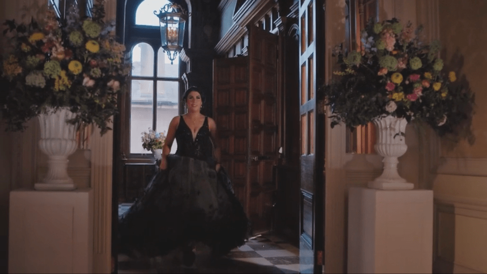 A woman in a black gown walks through an ornate doorway flanked by large floral arrangements.