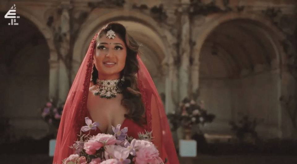 A bride in a red veil and heavily jeweled choker holds a bouquet of pink and purple flowers.