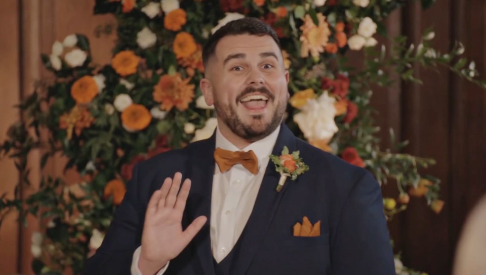 A groom in a blue suit with an orange bowtie and boutonnière gestures with his hand and sings, with a floral arrangement behind him.
