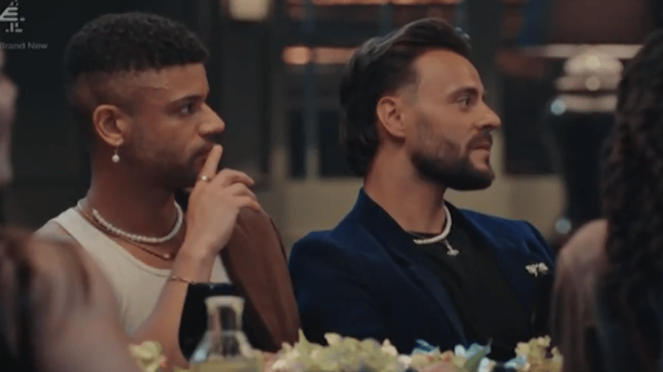 Two men at a table, one in a white tank top with pearl necklaces and an earring, the other in a black shirt and blue jacket with a necklace.