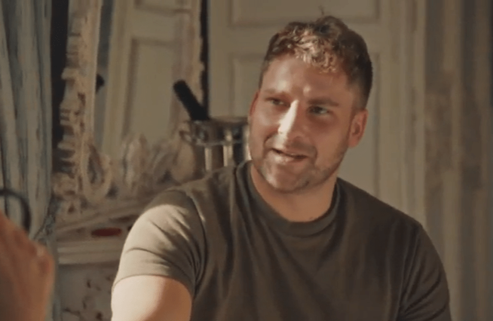 A light-skinned man with short brown hair and a beard, wearing a plain green t-shirt, looking away from the camera and smiling slightly.