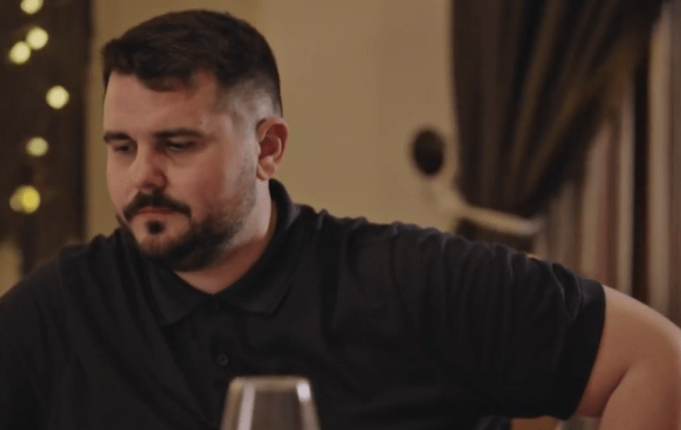 A man with dark hair and a beard, wearing a black polo shirt, looks down with a serious expression.