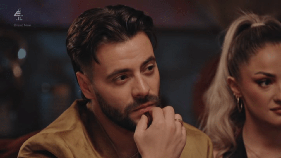 A man with a beard, wearing a gold jacket, looks to the right with a concerned expression, while a woman with blonde hair in a ponytail looks to the right next to him.