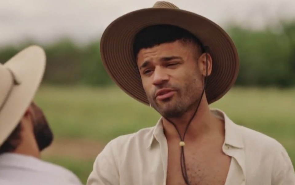 A man in a wide-brimmed hat speaks to another person, whose back is visible.