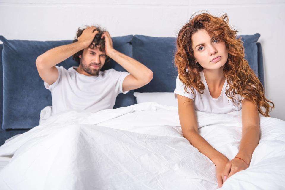 Young woman sitting on bed and looking at camera while man suffering from headache behind.