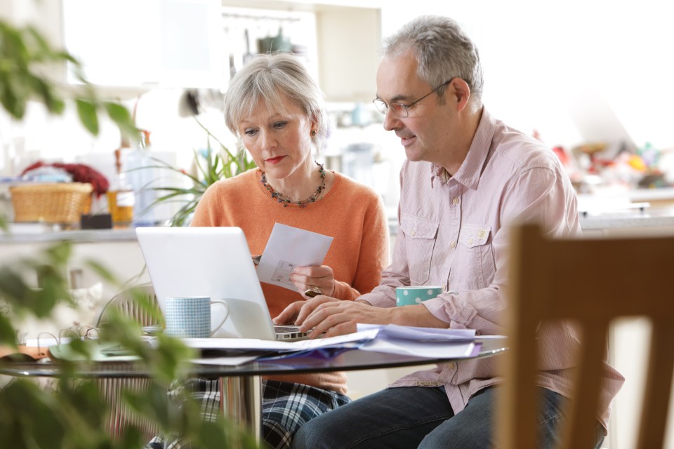 An older couple manages home finances, reviewing documents and using a laptop at a table.