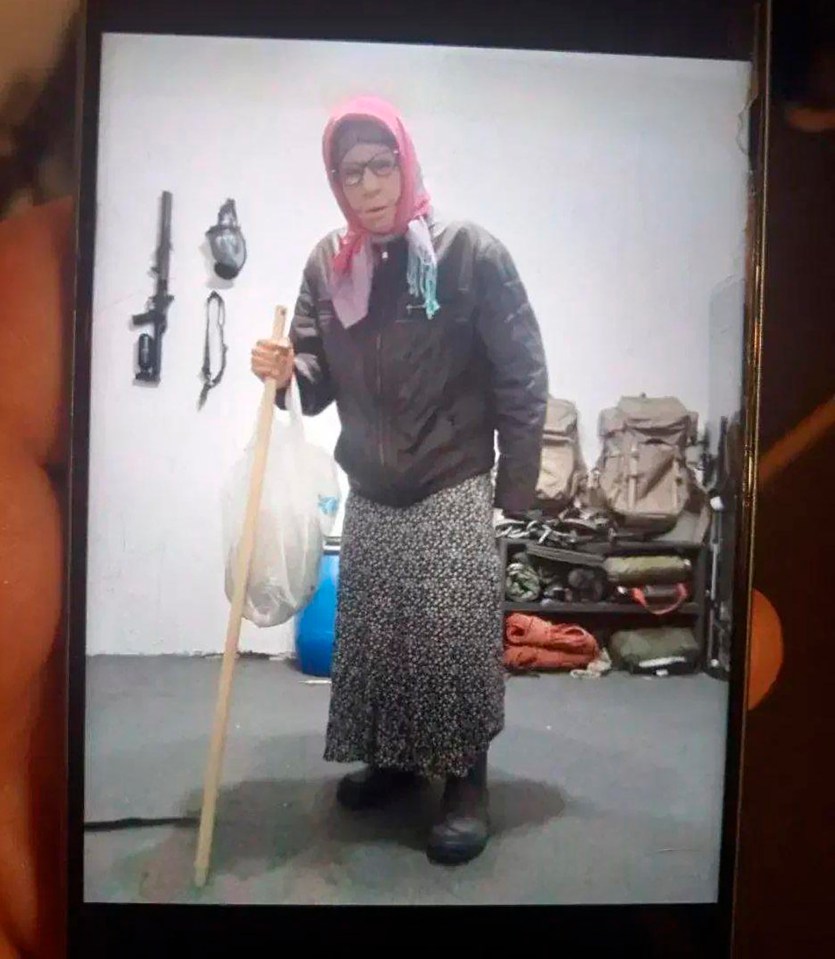 A man in women's clothing, including a pink headscarf and a patterned skirt, holding a stick and a white plastic bag, standing in a room with military equipment.
